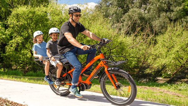 A man rides an orange electric cargo bike with two children seated on the rear. All three are wearing helmets and smiling as they ride along a paved path with greenery in the background.